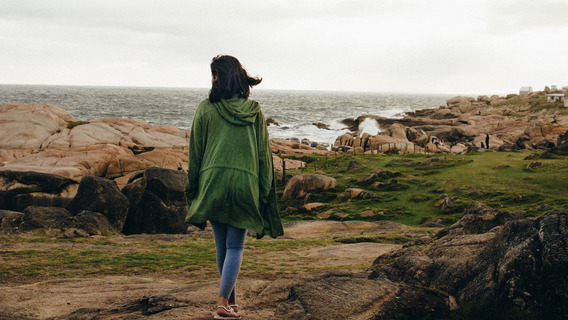 Eine Person in einem grünen Mantel läuft, uns den Rücken zugekehrt, den felsigen Strand von Cabo Polonio in Uruguay entlang, auf den tosende Wellen treffen.