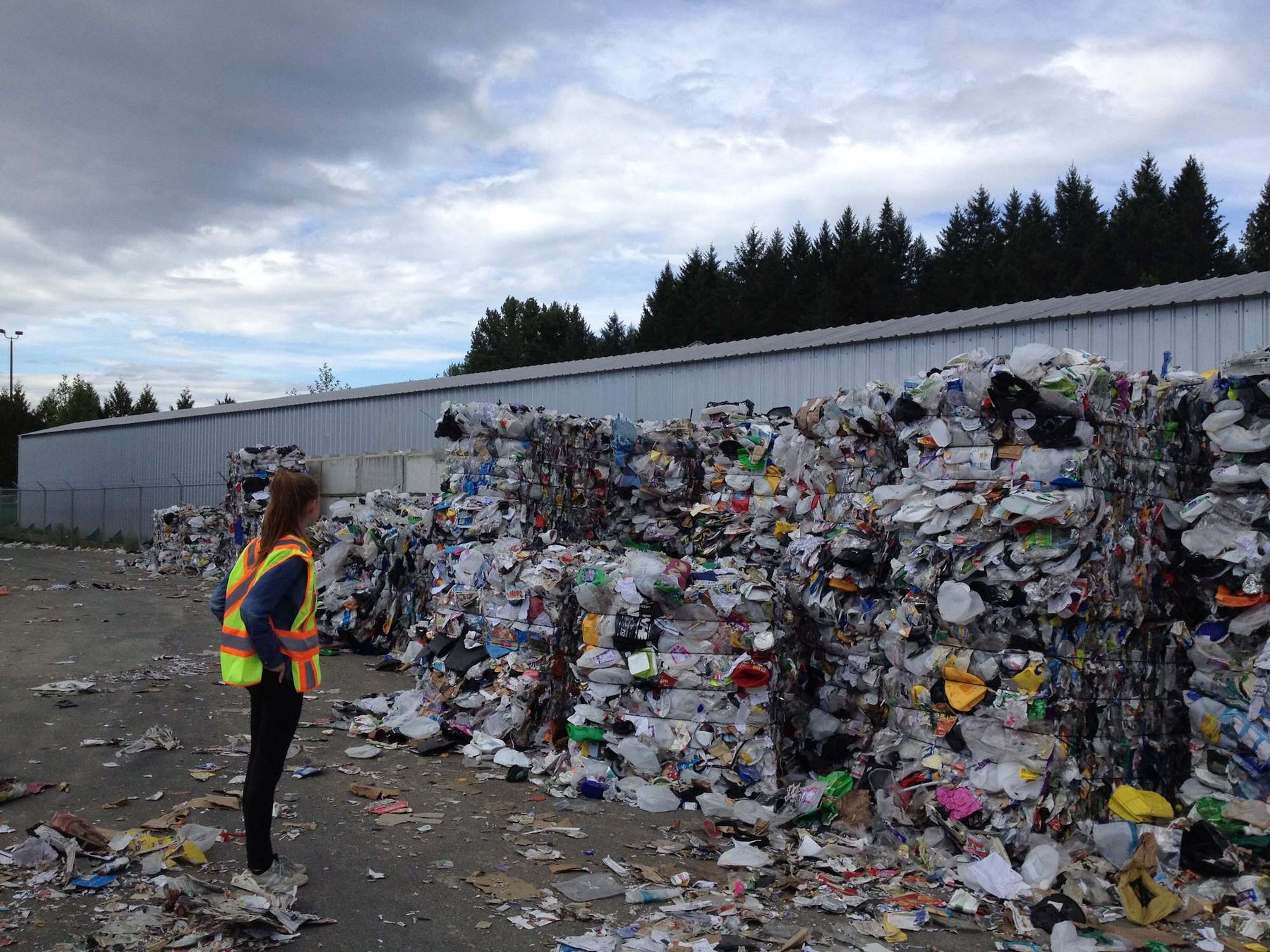 Eine Frau steht vor zu vielen großen Bündeln zusammengeschnürtem Verpackungsmüll auf dem Hof einer Recyclinganlage