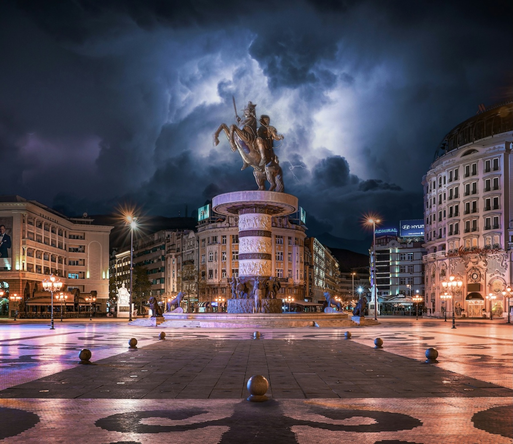 Eine monumentale Reiterstatue thront auf einem beleuchteten Brunnen in der Mitte eines Platzes in Skopje, während sich dramatische dunkle Gewitterwolken am nächtlichen Himmel auftürmen. Die umliegenden Gebäude und Straßenlaternen tauchen den menschenleeren Platz in warmes Licht, das einen starken Kontrast zur bedrohlichen Atmosphäre bildet.