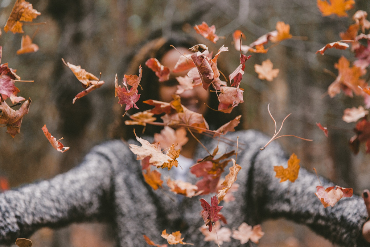 Eine Frau in grauem Mantel wirft buntes Herbstlaub vor sich in die Höhe. Sie lächelt dabei fröhlich.