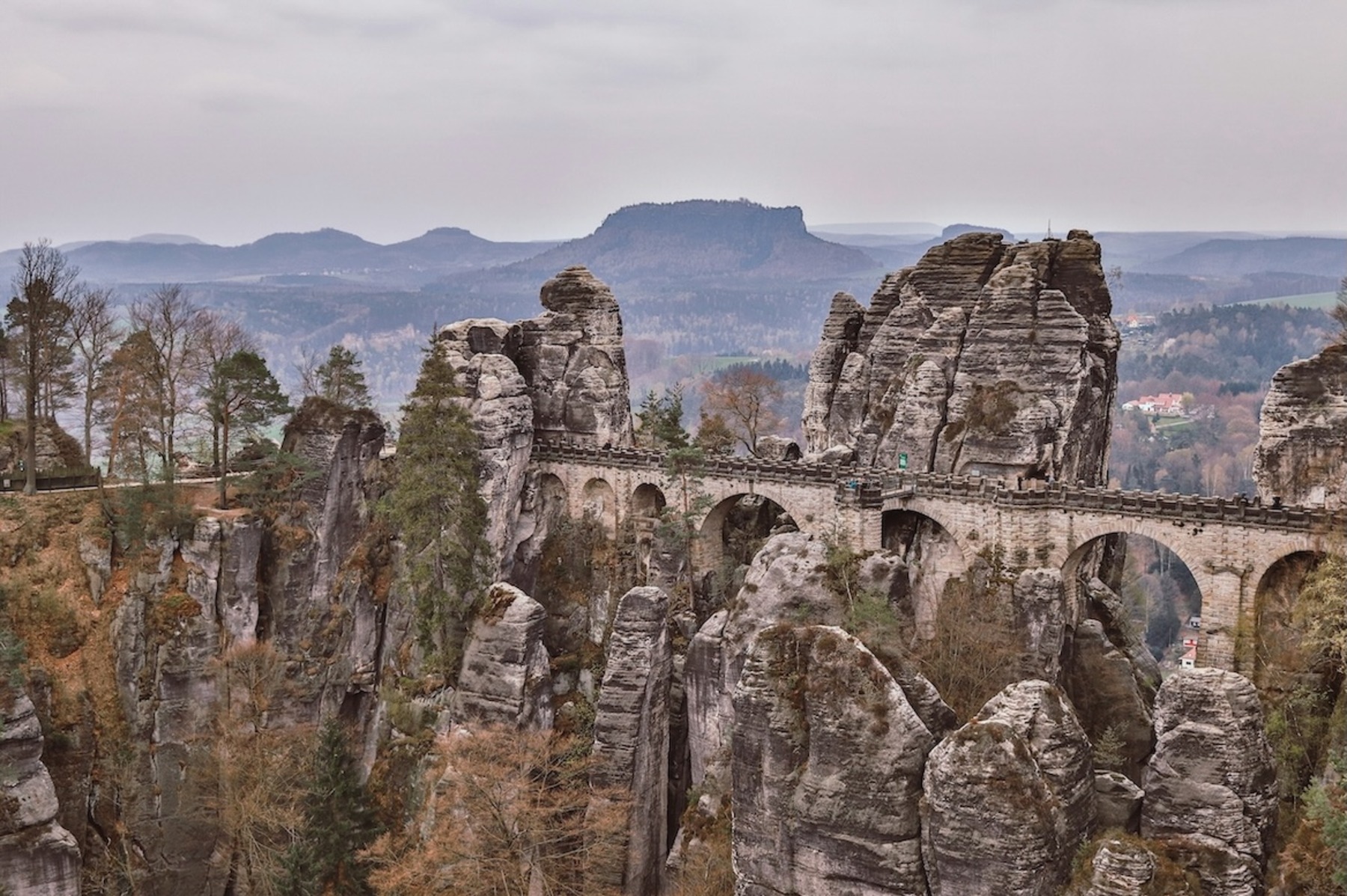 Zu sehen ist eine historische Brücke, die sich zwischen Felsformationen aufspannt, im Hintergrund erstrecken sich Wälder und Berge soweit das Auge reicht