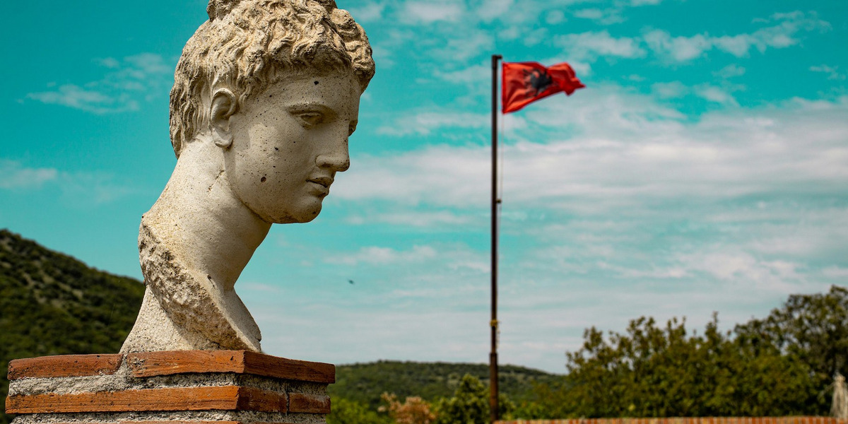 : Zu sehen sind der Kopf einer antiken Statue vor einer mediterranen Hügellandschaft im Nationalpark Butrint. Im Hintergrund weht die albanische Flagge.