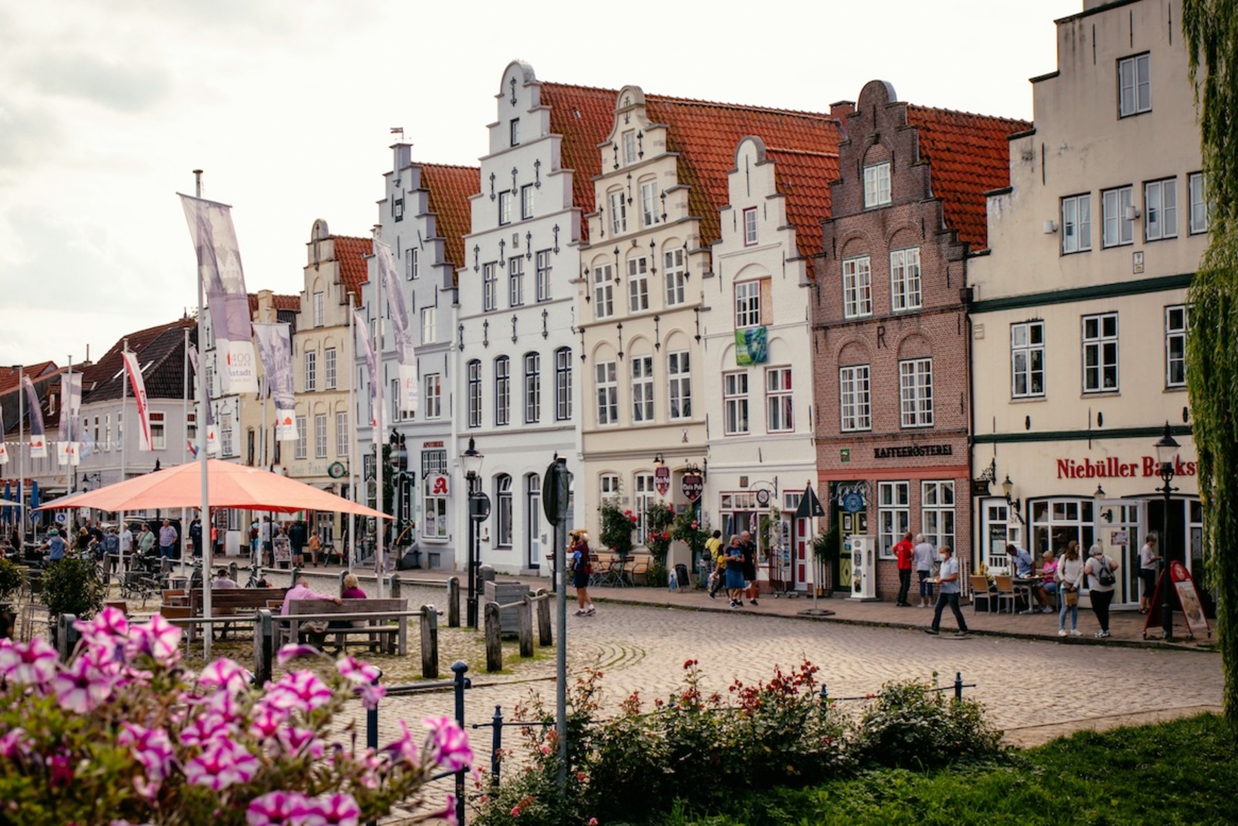 Eine Reihe von historischen Häusern mit Treppengiebeln und roten Dächern in Friedrichstadt, Schleswig-Holstein. Auf der gepflasterten Straße spazieren Menschen oder sitzen unter einem orangefarbenen Sonnenschirm. Im Vordergrund blühen rosa und lilafarbene Blumen.