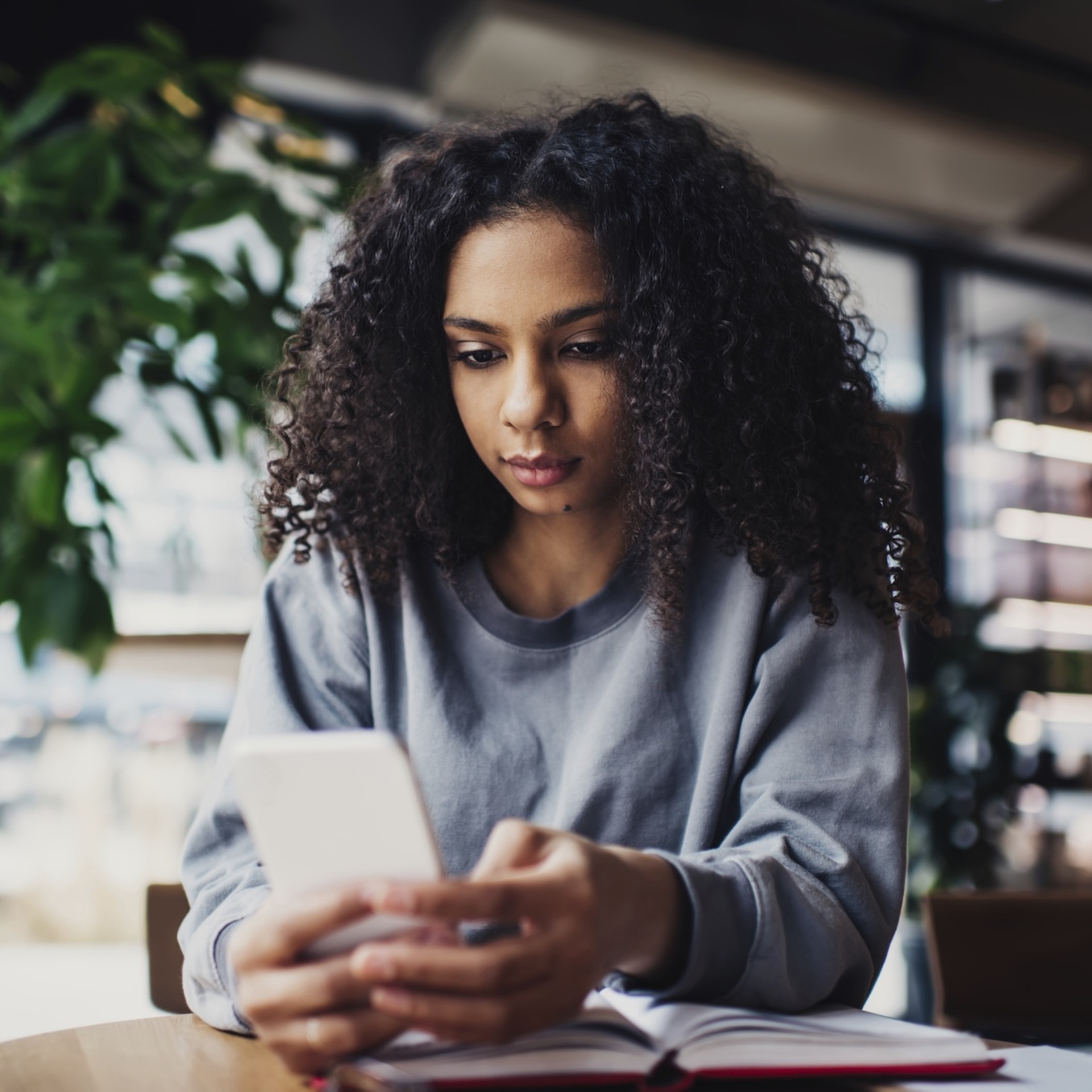 Eine junge Frau mit lockigem Haar sitzt in einem Café und blickt auf ihr Smartphone. Vor ihr liegt ein geöffnetes Notizbuch auf dem Tisch. Im Hintergrund sind Pflanzen und Regale zu sehen.