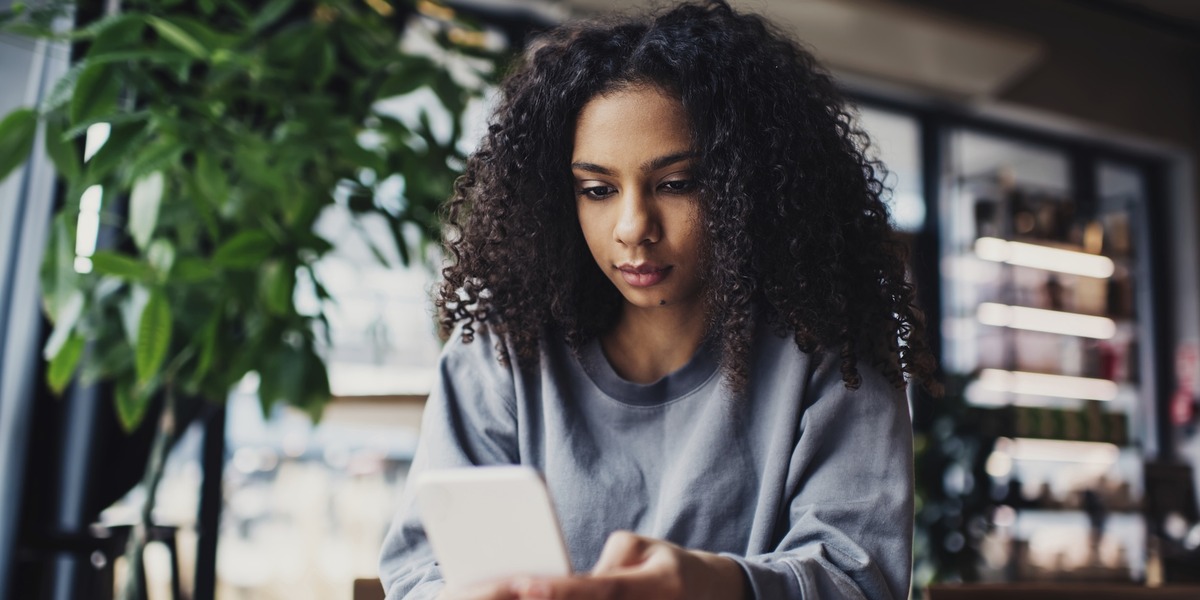 Eine junge Frau mit lockigem Haar sitzt in einem Café und blickt auf ihr Smartphone. Vor ihr liegt ein geöffnetes Notizbuch auf dem Tisch. Im Hintergrund sind Pflanzen und Regale zu sehen.