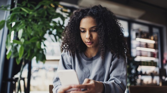 Eine junge Frau mit lockigem Haar sitzt in einem Café und blickt auf ihr Smartphone. Vor ihr liegt ein geöffnetes Notizbuch auf dem Tisch. Im Hintergrund sind Pflanzen und Regale zu sehen.