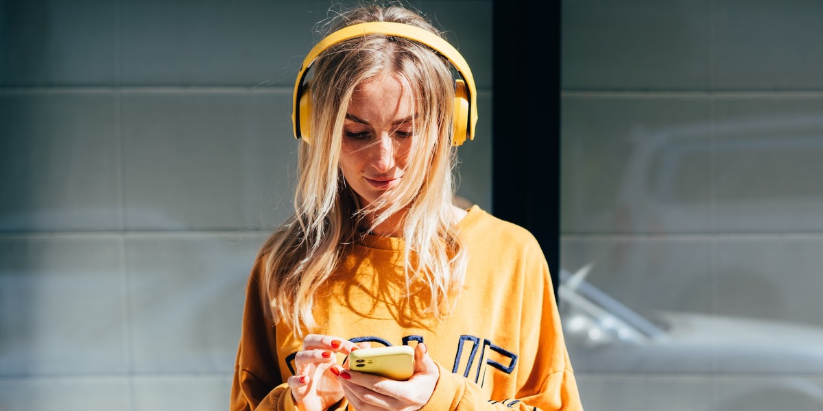Junge Frau mit gelben Over-Ear-Kopfhörern und gelbem Oversized-Sweatshirt, die auf ihr gelbes Smartphone schaut. Sie steht vor einer verglasten Fassade, im Hintergrund ist ein silberfarbenes Auto zu erkennen.