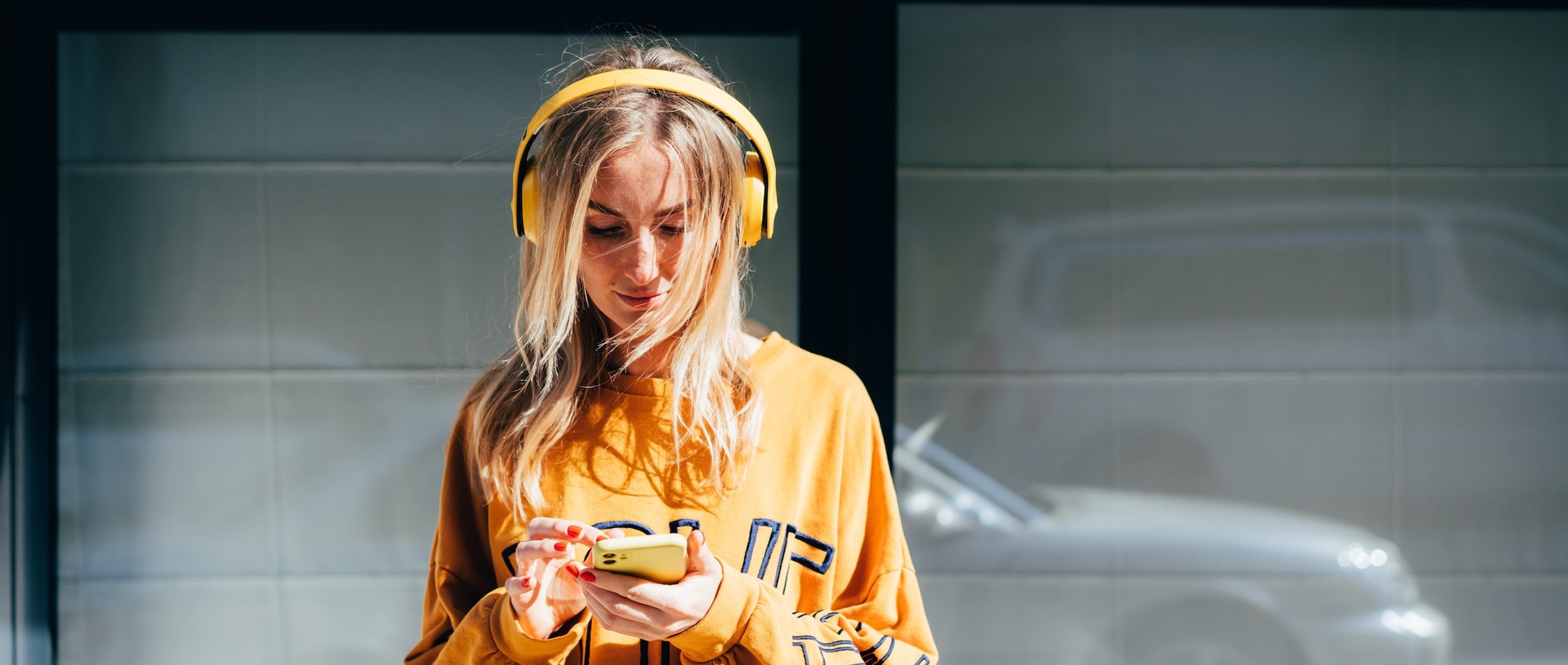 Junge Frau mit gelben Over-Ear-Kopfhörern und gelbem Oversized-Sweatshirt, die auf ihr gelbes Smartphone schaut. Sie steht vor einer verglasten Fassade, im Hintergrund ist ein silberfarbenes Auto zu erkennen.