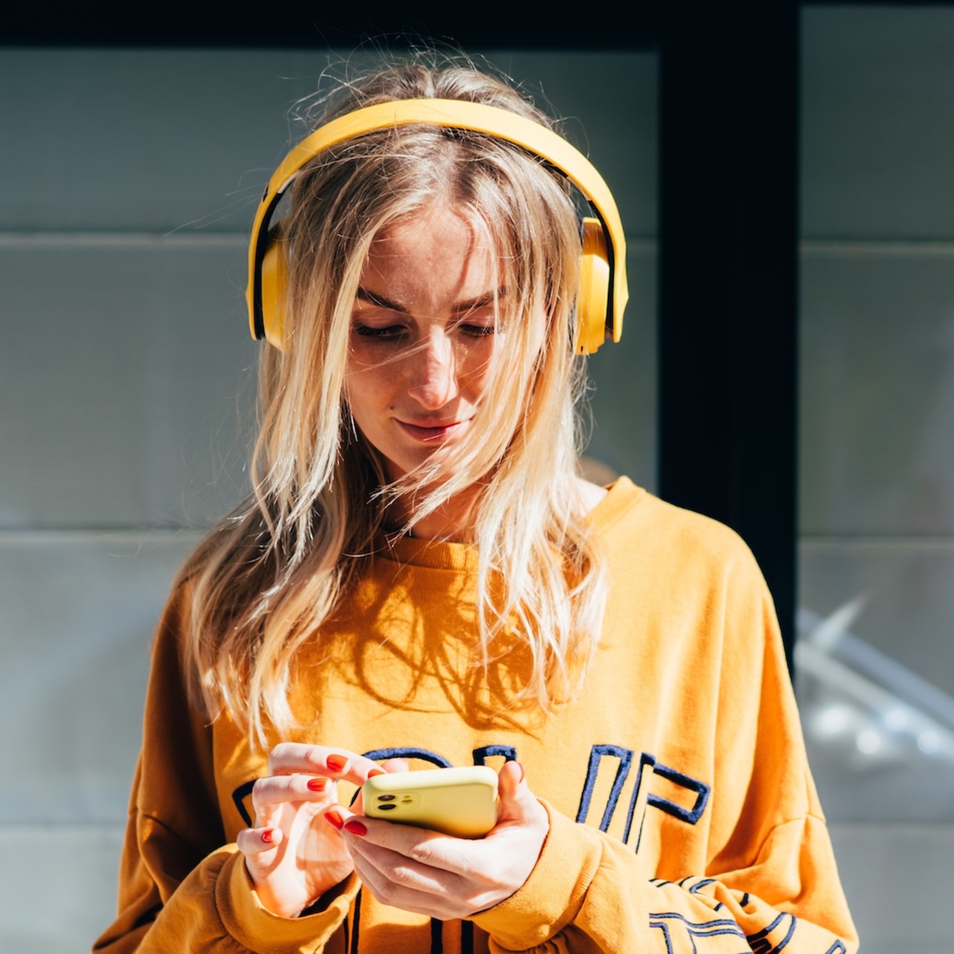 Junge Frau mit gelben Over-Ear-Kopfhörern und gelbem Oversized-Sweatshirt, die auf ihr gelbes Smartphone schaut. Sie steht vor einer verglasten Fassade, im Hintergrund ist ein silberfarbenes Auto zu erkennen.