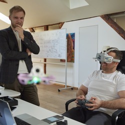 Zwei Männer in einem modernen Büro. Der sitzende Mann im Vordergrund trägt eine Virtual-Reality-Brille und einen Controller in den Händen, während er eine Drohne steuert, die vor ihm schwebt. Der andere Mann steht neben ihm und beobachtet.