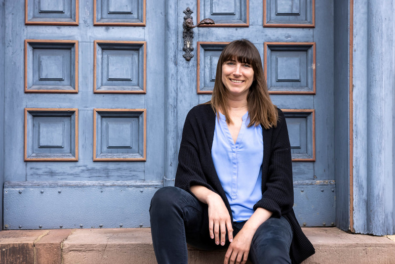 Das Foto zeigt Sarah Ackermann, die auf einer Treppe vor einer blau lackierten Tür sitzt. Sie schaut freundlich in die Kamera und lächelt.