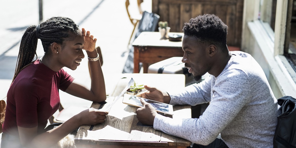 Ein junges Paar sitzt an einem Tisch vor einem Café und unterhält sich. Die Frau trägt einen weinroten Pullover und lächelt, während der Mann ihr zugewandt in einem hellen Hemd am Tisch sitzt.
