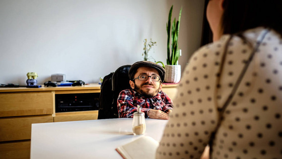 Ein Mann mit Brille und Kappe sitzt gegenüber von einer anderen Person an einem Tisch.