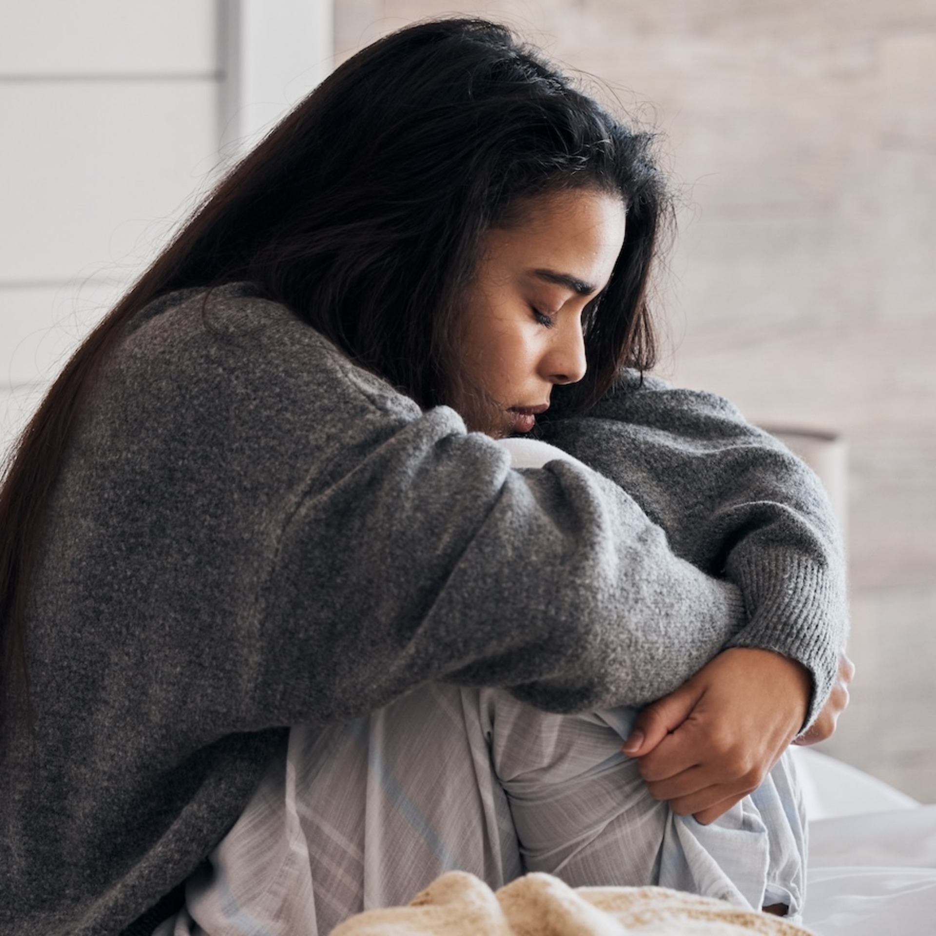 Eine junge Frau sitzt nachdenklich auf einem Bett mit weißer Bettwäsche und zieht ihre Knie an sich. Sie trägt einen grauen Pullover und blickt nach unten, während sie in sich gekehrt wirkt.