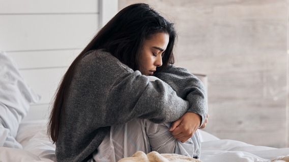 Eine junge Frau sitzt nachdenklich auf einem Bett mit weißer Bettwäsche und zieht ihre Knie an sich. Sie trägt einen grauen Pullover und blickt nach unten, während sie in sich gekehrt wirkt.