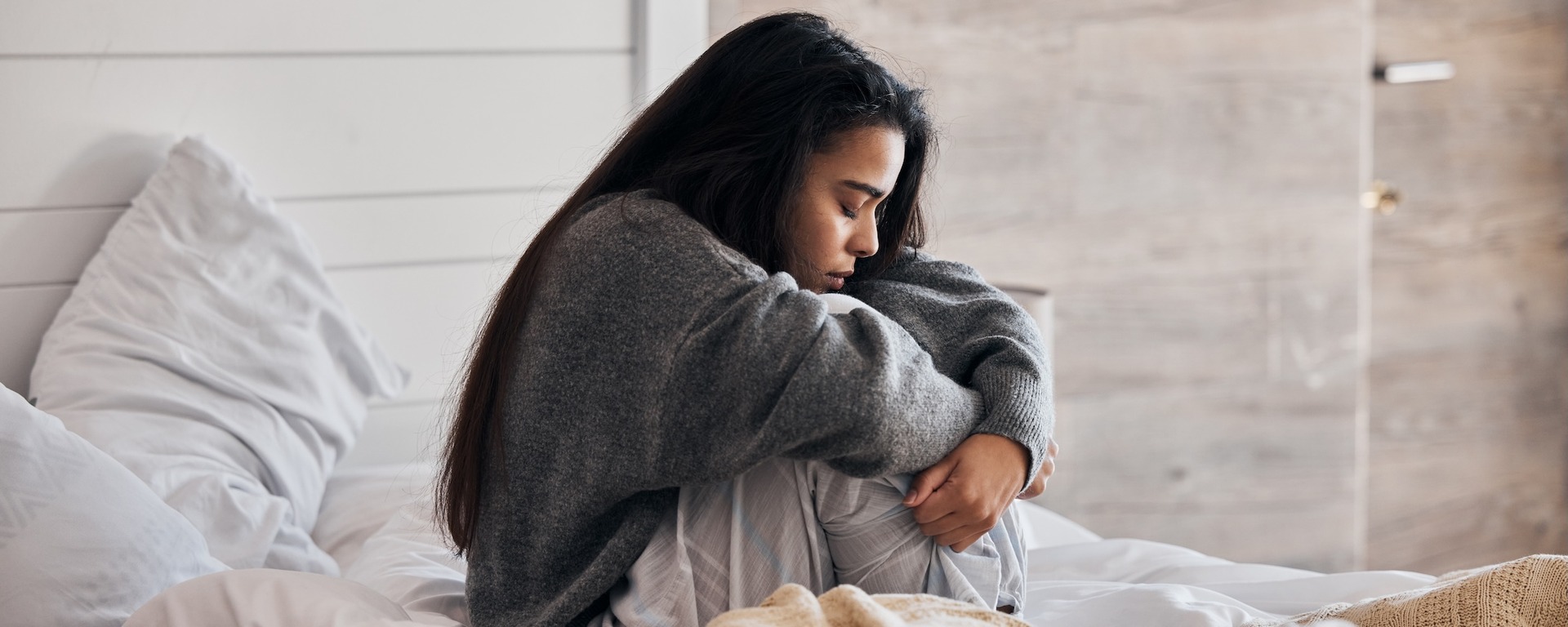 Eine junge Frau sitzt nachdenklich auf einem Bett mit weißer Bettwäsche und zieht ihre Knie an sich. Sie trägt einen grauen Pullover und blickt nach unten, während sie in sich gekehrt wirkt.