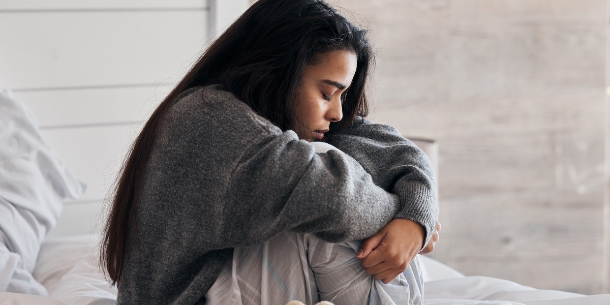 Eine junge Frau sitzt nachdenklich auf einem Bett mit weißer Bettwäsche und zieht ihre Knie an sich. Sie trägt einen grauen Pullover und blickt nach unten, während sie in sich gekehrt wirkt.