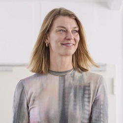 Eine lächelnde Frau steht in einem hellen Raum und blickt zur Seite. Im Hintergrund sind eine große Zimmerpflanze, aufgehängte Jacken, mehrere Kartons und ein Bildschirm an der Wand zu sehen.