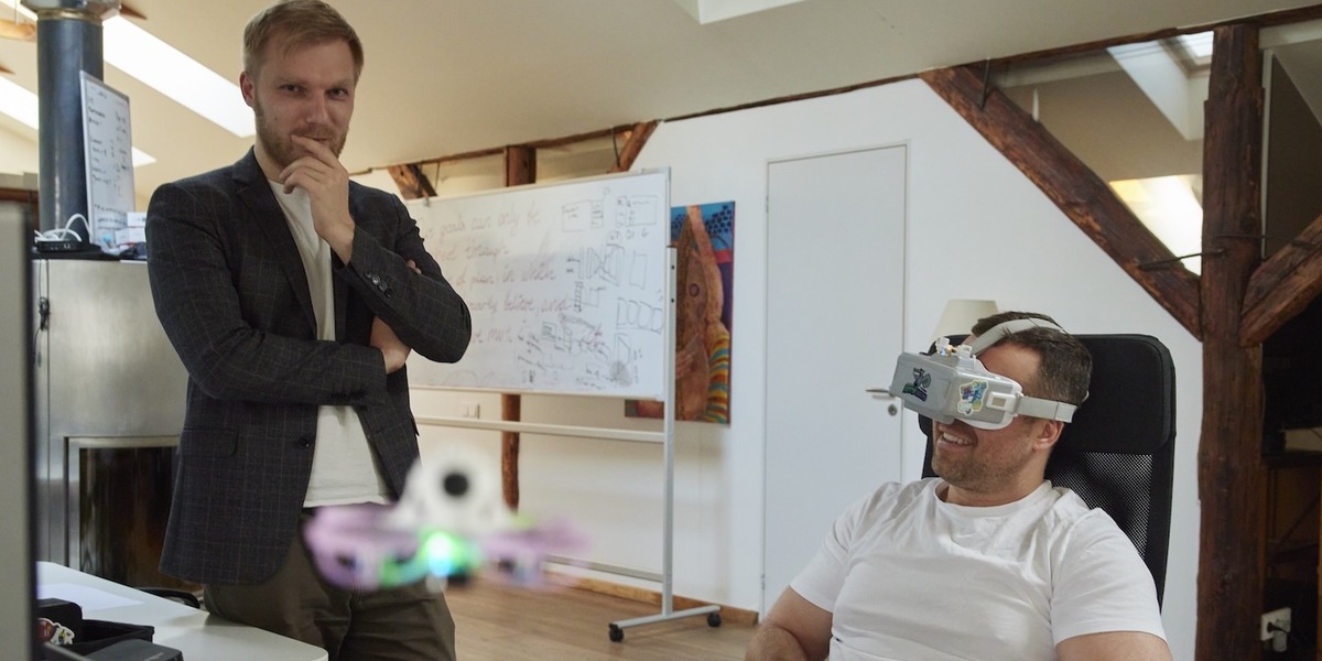 Zwei Männer in einem modernen Büro. Der sitzende Mann im Vordergrund trägt eine Virtual-Reality-Brille und einen Controller in den Händen, während er eine Drohne steuert, die vor ihm schwebt. Der andere Mann steht neben ihm und beobachtet.