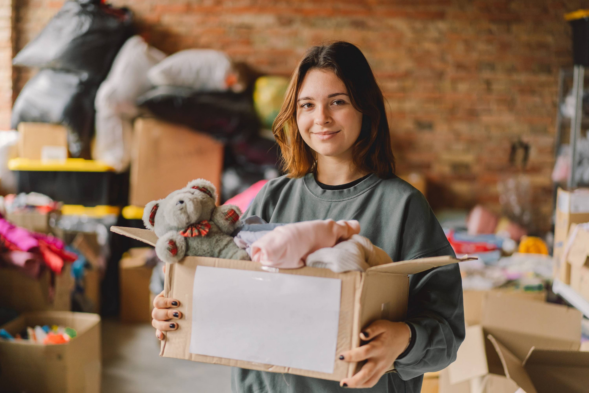 Eine Jugendliche hält einen Karton mit einem Teddybären und Kleidung. Im Hintergrund weitere Kartons mit Sachspenden.