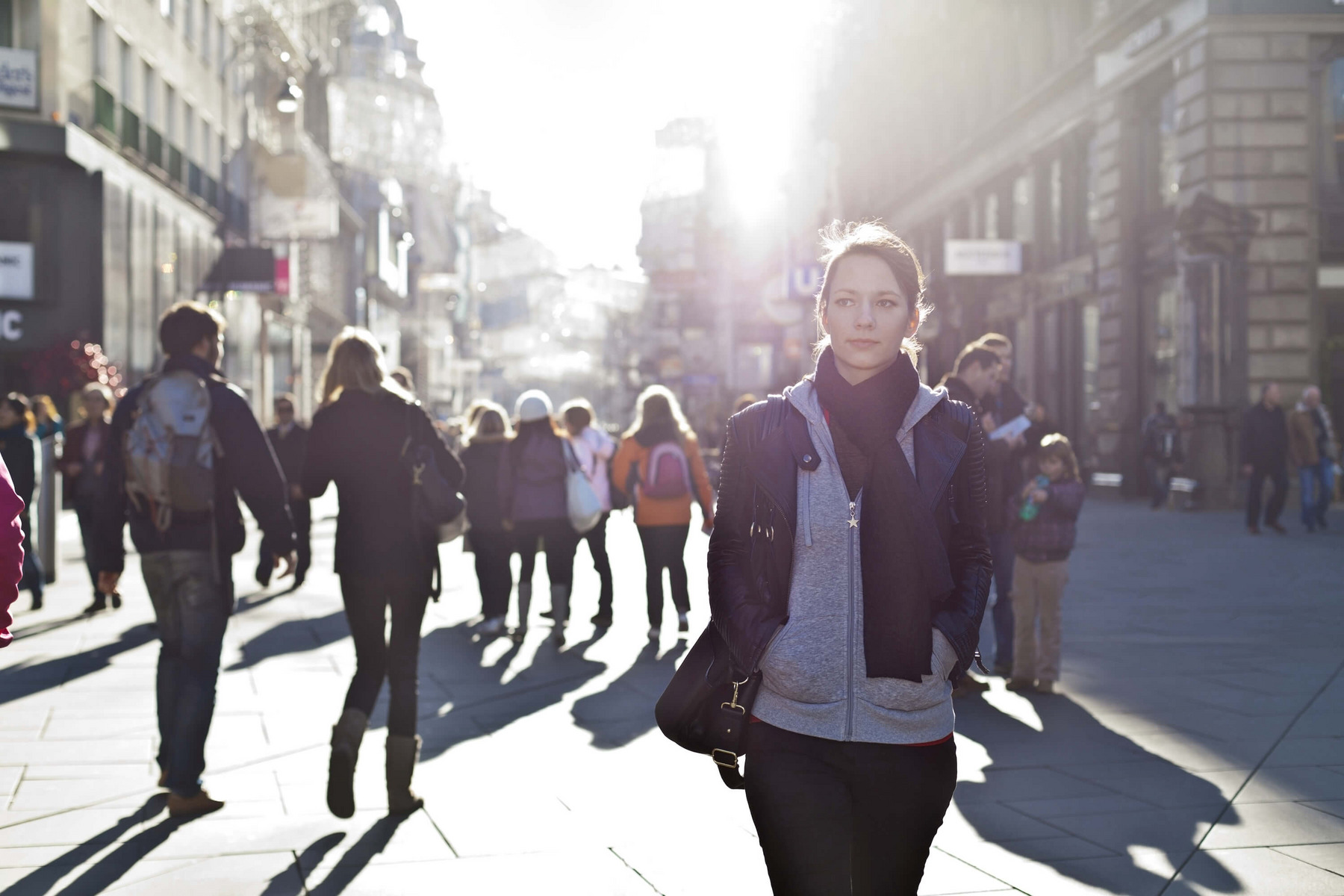 Eine Frau mit vielen anderen Passant:innen im Hintergrund geht durch eine Fußgänger:innenzone einer Innenstadt. Die Straße ist umgeben von urbaner Bebauung, die Sonne scheint hell im Hintergrund.