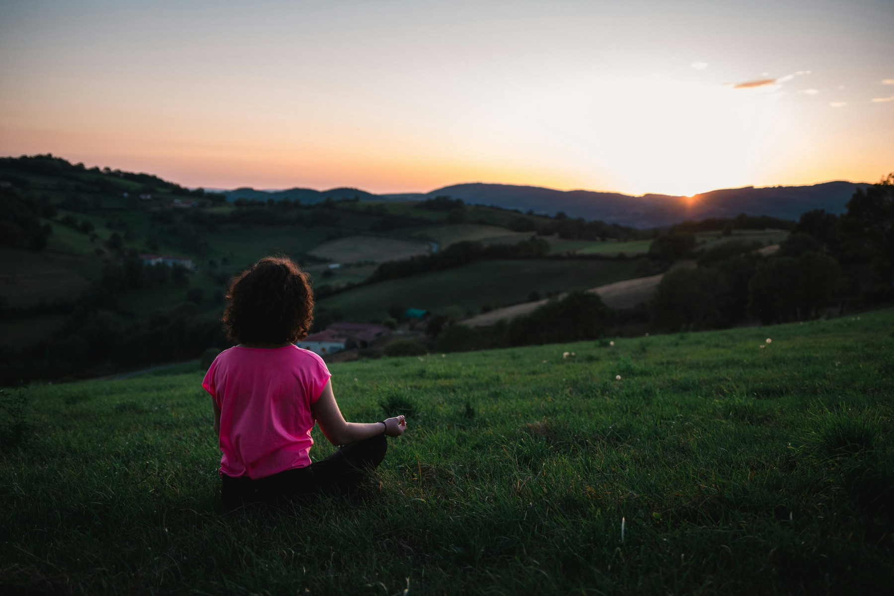 Eine Frau im pinken T-Shirt sitzt in Meditationshaltung auf einem grünen Hügel, im Hintergrund geht die Sonne unter.