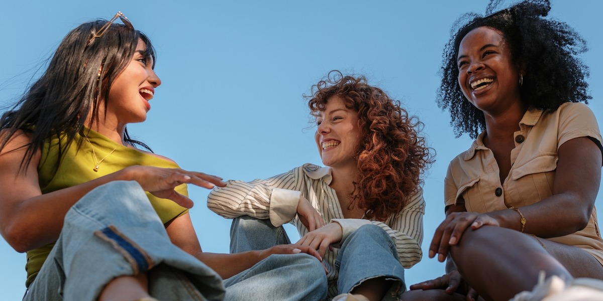 Drei junge Frauen sitzen gemeinsam lachend auf einer Wiese im Sonnenschein.