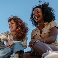 Drei junge Frauen sitzen gemeinsam lachend auf einer Wiese im Sonnenschein.