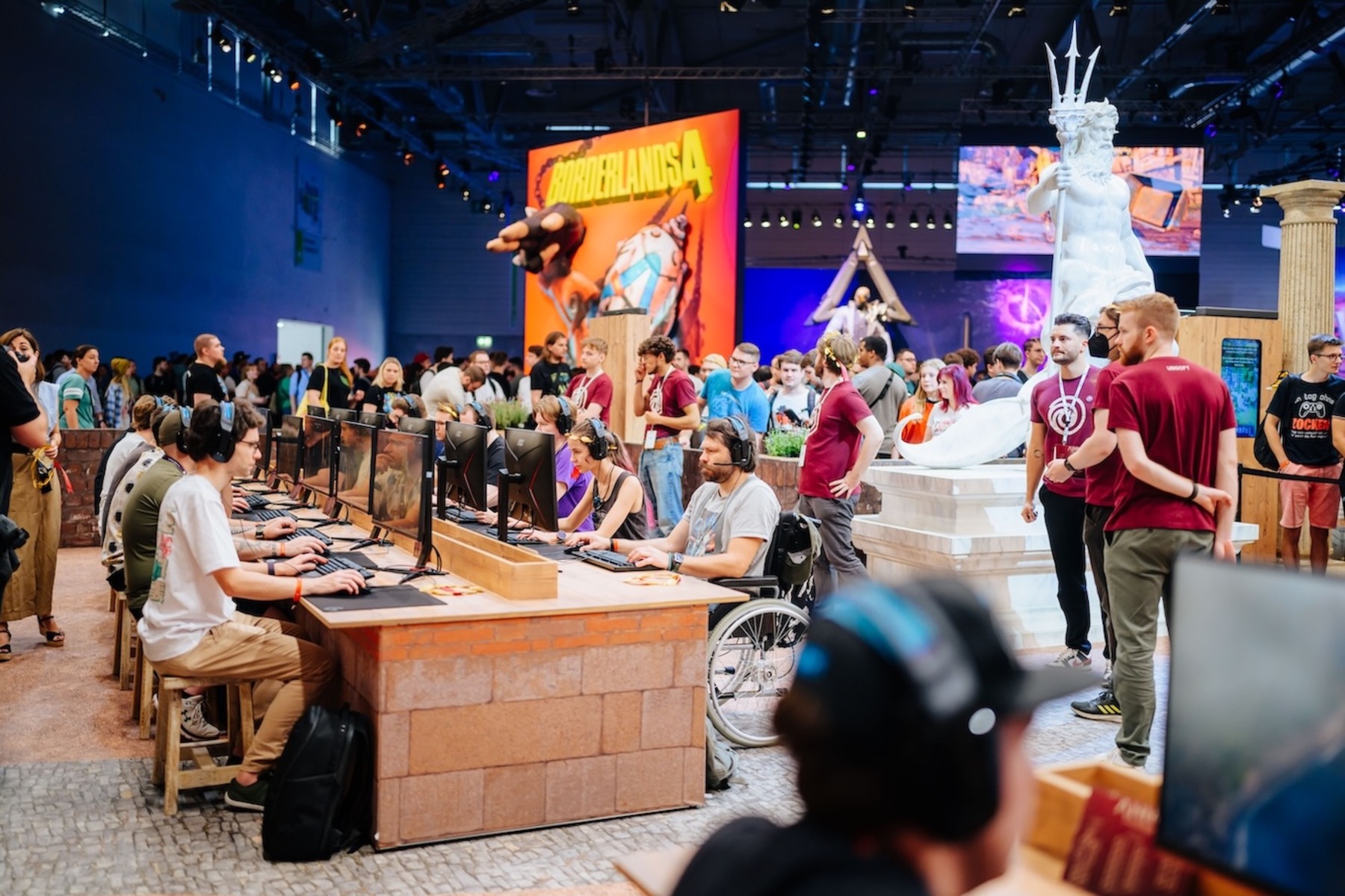 Das Bild zeigt die gamescom, eine große Videospiel-Messe mit vielen Besucher:innen. Im Bild sitzen zahlreiche Menschen an großen Tischen und spielen an Computern.