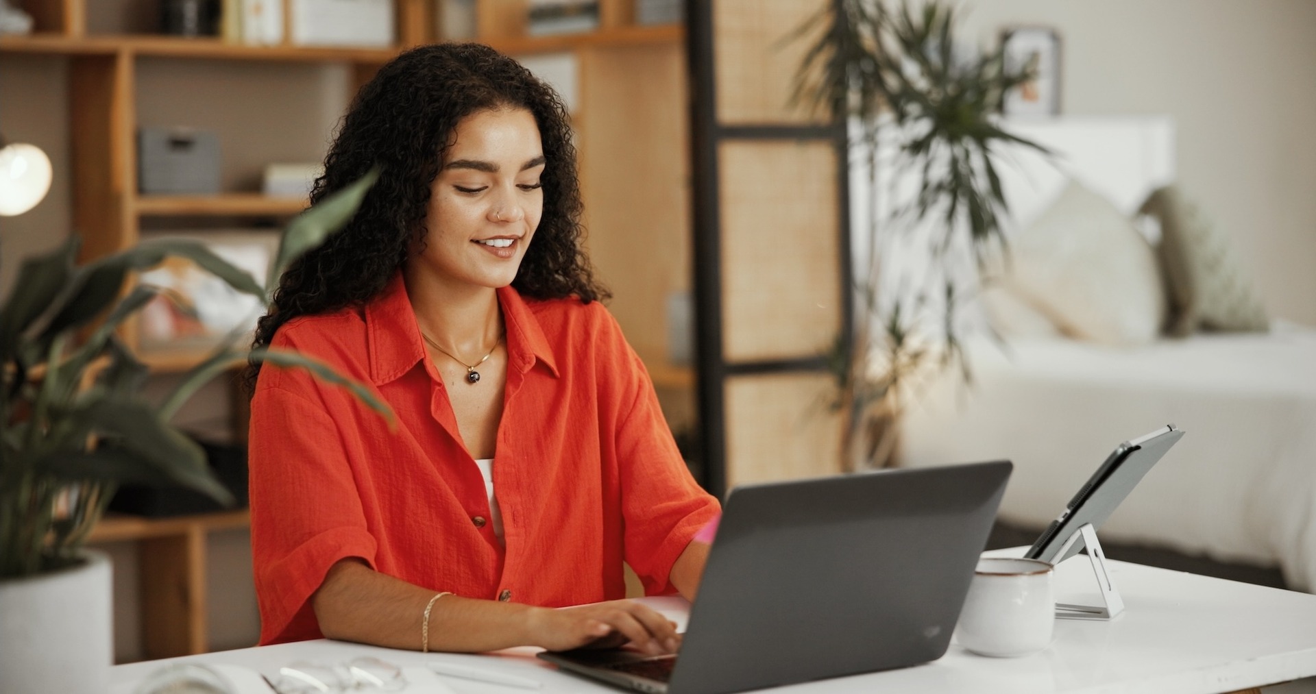 Eine Frau mit lockigen Haaren sitzt an einem Tisch und arbeitet lächelnd an einem Laptop. Im Hintergrund sind Pflanzen und ein Regal in einer wohnlichen Umgebung zu sehen.