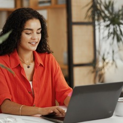 Eine Frau mit lockigen Haaren sitzt an einem Tisch und arbeitet lächelnd an einem Laptop. Im Hintergrund sind Pflanzen und ein Regal in einer wohnlichen Umgebung zu sehen.
