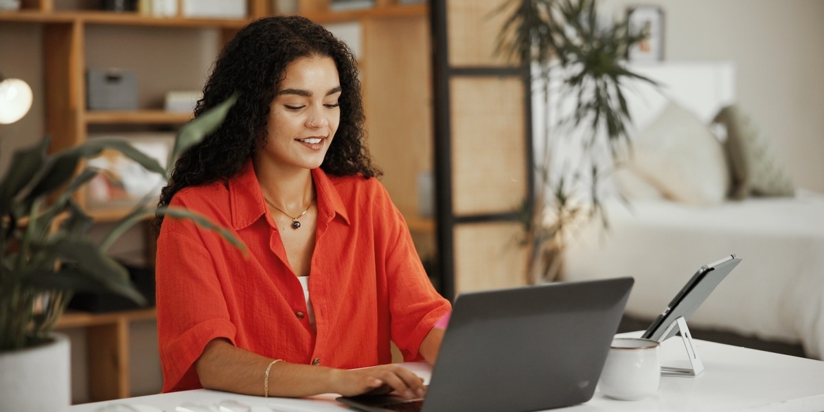 Eine Frau mit lockigen Haaren sitzt an einem Tisch und arbeitet lächelnd an einem Laptop. Im Hintergrund sind Pflanzen und ein Regal in einer wohnlichen Umgebung zu sehen.