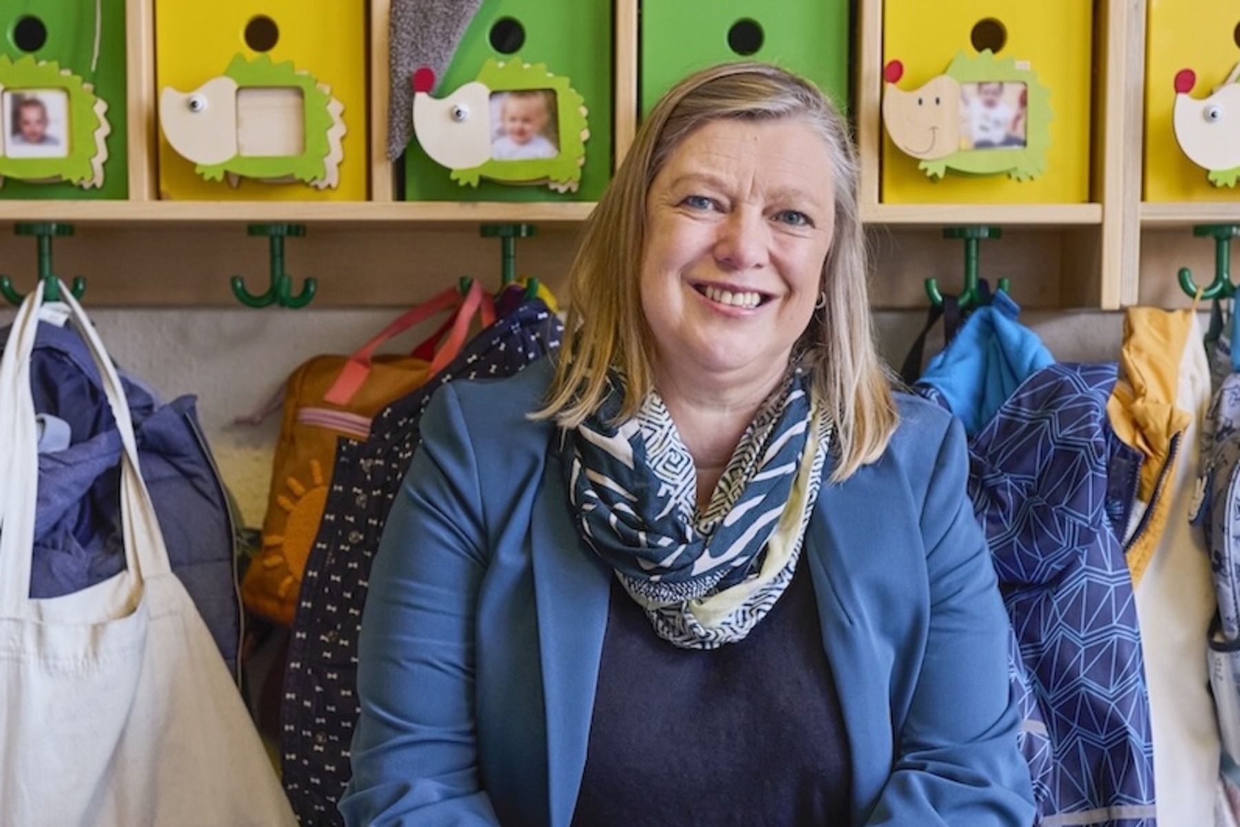 Eine lächelnde Frau sitzt auf einer Holzbank in einer Kindergarderobe. Hinter ihr befinden sich offene Fächer mit bunten Aufbewahrungsboxen, Kinderkleidung, Schuhen, Taschen und Rucksäcken.