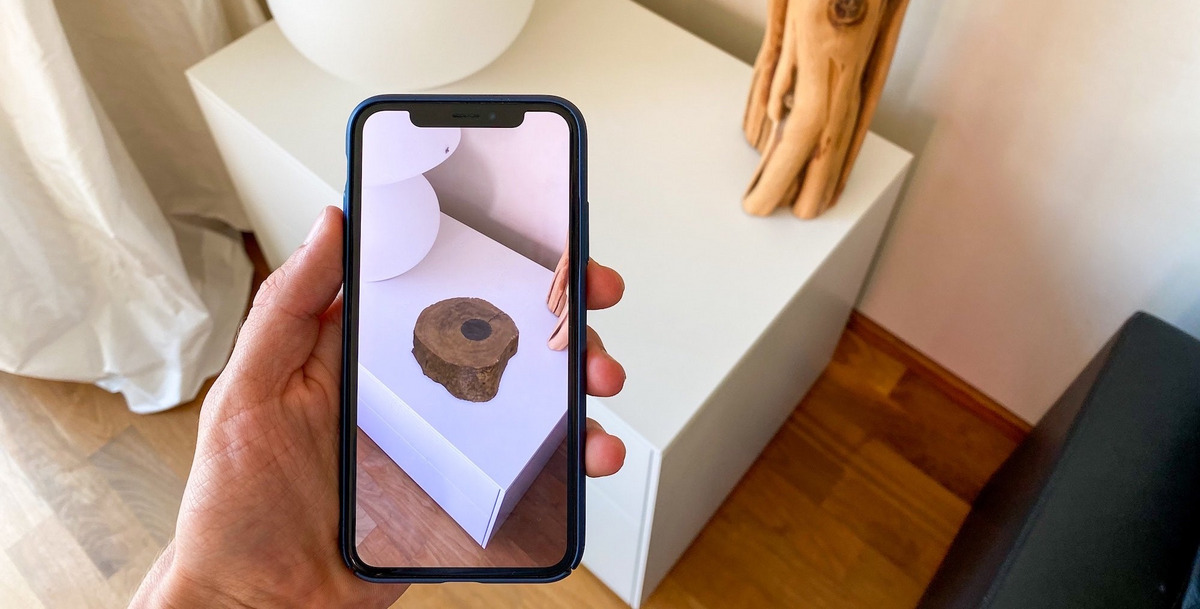 Das Foto zeigt eine Hand, die ein Smartphone vor eine Kommode hält. Auf der Oberfläche erscheint mittels Augmented Reality ein Designobjekt in Form einer Holzskulptur.