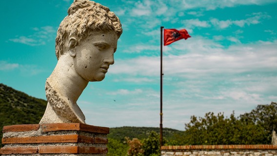 : Zu sehen sind der Kopf einer antiken Statue vor einer mediterranen Hügellandschaft im Nationalpark Butrint. Im Hintergrund weht die albanische Flagge.