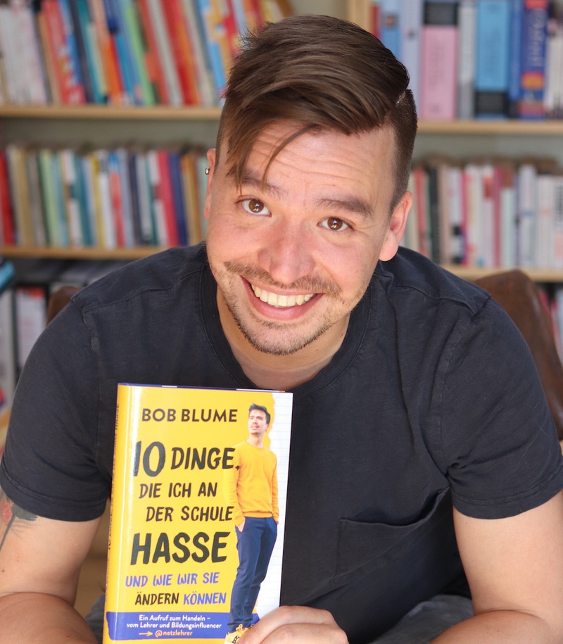 Bob Blume sitzt vor einem Bücherregal, in der Hand hält er sein eigenes Buch mit dem Titel „10 Dinge, die ich an der Schule hasse und wie wir sie ändern können“.