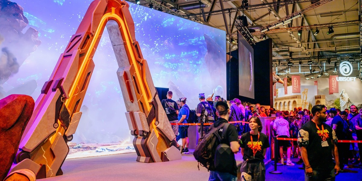Eine große, orange beleuchtete Spielzeug-Spitzhacke steht als Ausstellungsstück auf einer Gaming-Messe, während Besucher vor einer Videowand mit Fantasy-Landschaft vorbeigehen. Im Hintergrund sind Messestände verschiedener Aussteller wie Ubisoft zu sehen.