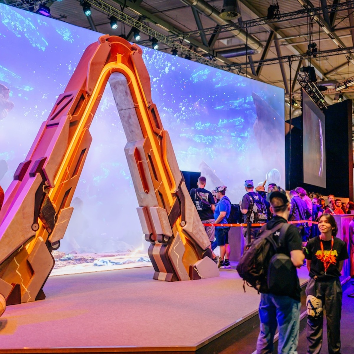 Eine große, orange beleuchtete Spielzeug-Spitzhacke steht als Ausstellungsstück auf einer Gaming-Messe, während Besucher vor einer Videowand mit Fantasy-Landschaft vorbeigehen. Im Hintergrund sind Messestände verschiedener Aussteller wie Ubisoft zu sehen.