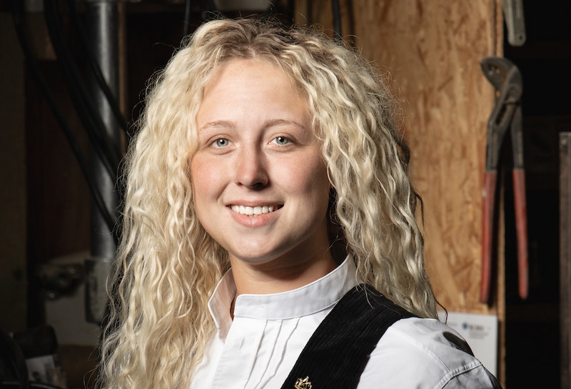 Eine junge Frau mit langen blonden Locken trägt eine traditionelle Tracht mit schwarzer Lederweste und weißer Bluse und lächelt freundlich in die Kamera.
