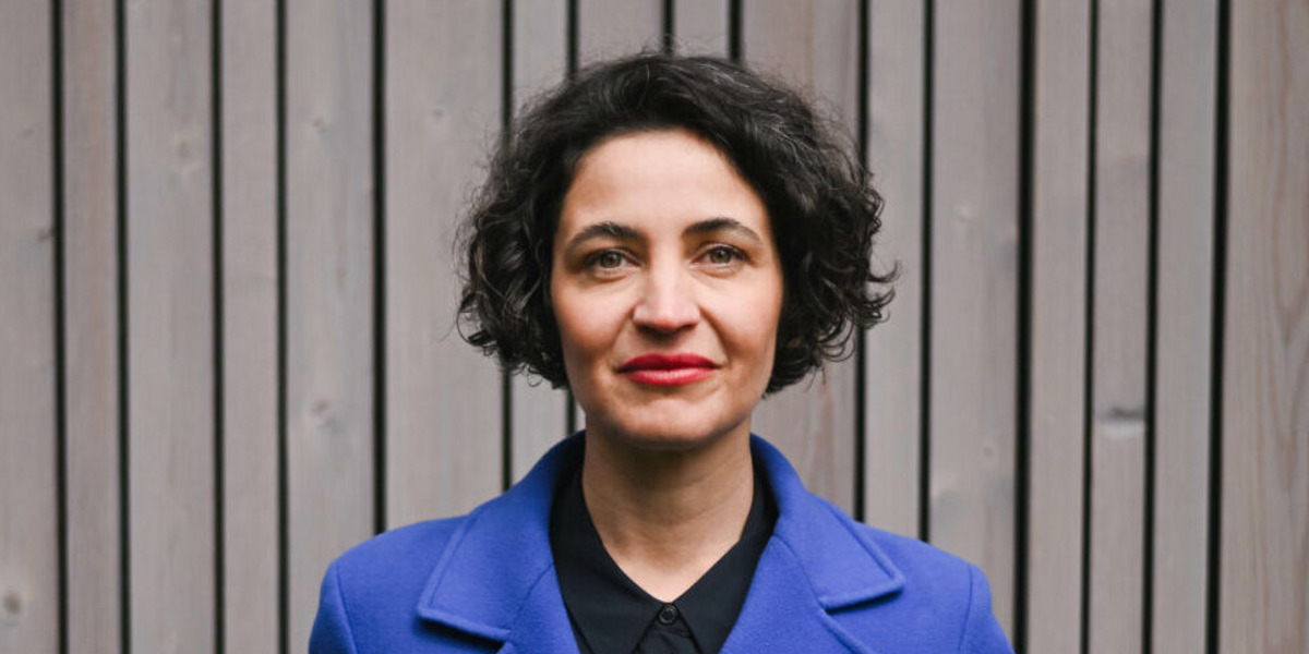 Aufnahme von Anna-Lena von Hodenberg vor einem Holzzaun. Sie trägt einen strahlendblauen Mantel und schaut in die Kamera, die Hände locker in die Taschen gesteckt.