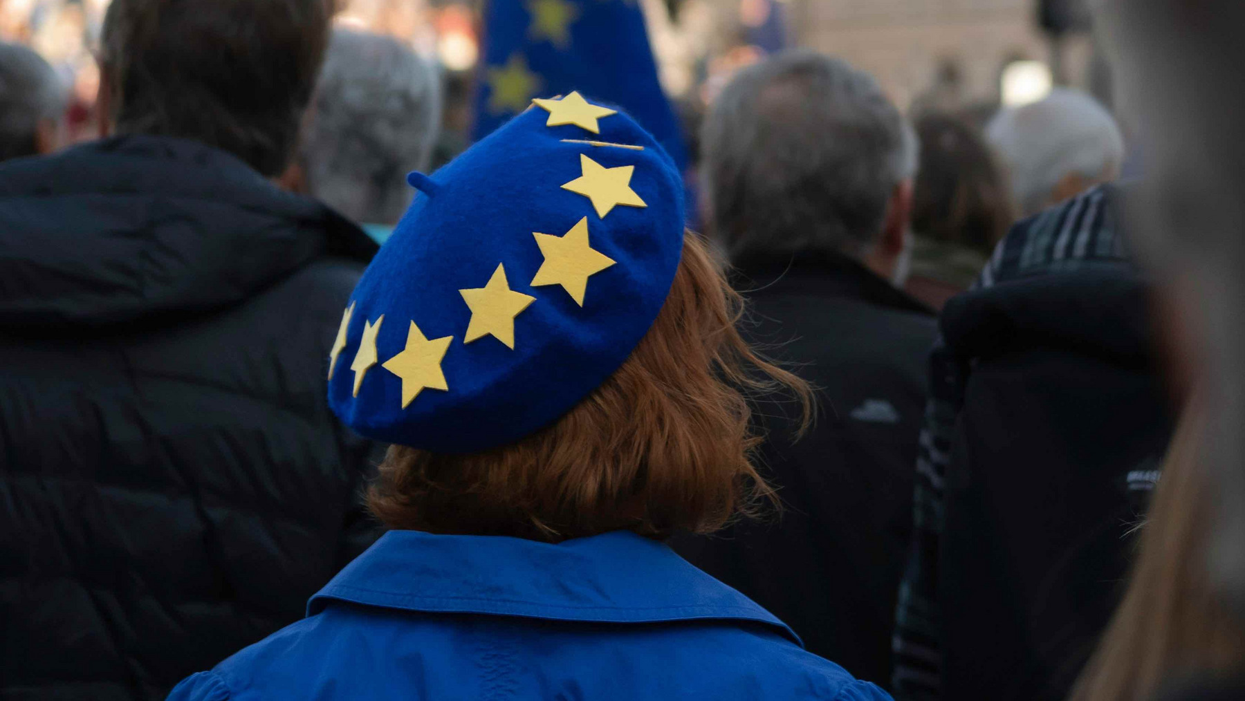europa-demo-baskenmuetze-change-magazin Das Bild zeigt eine Menschenmenge bei einer Veranstaltung im Freien. Im Vordergrund ist der Rücken einer Person zu sehen, die eine blaue Baskenmütze mit gelben Sternen, ähnlich der Europaflagge, trägt. Die Person trägt ebenfalls eine blaue Jacke. Im Hintergrund sind unscharf Gebäude und eine Säule zu erkennen. Die Veranstaltung scheint in einer Stadt, möglicherweise auf einem öffentlichen Platz, stattzufinden.