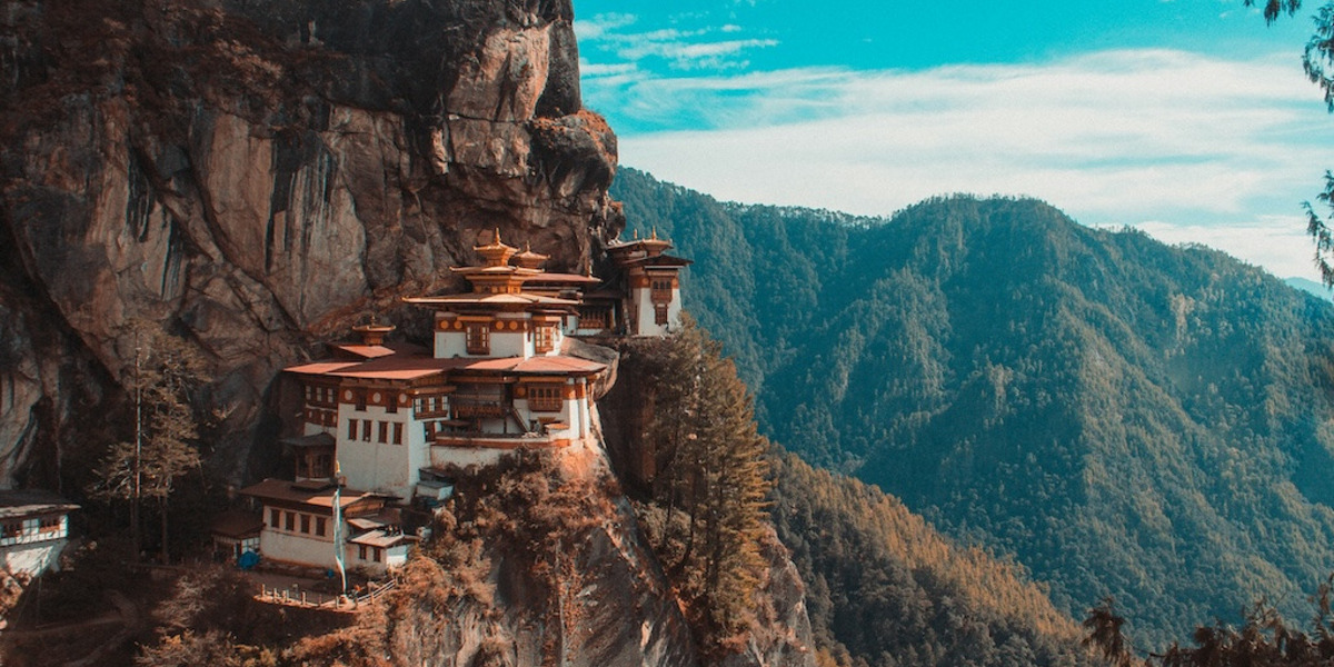 Das Taktshang-Kloster in Bhutan: Zu sehen sind eine Reihe von Tempeln, die sich an einen steilen Berghang schmiegen. Im Hintergrund sind weitere bewaldete Berghänge sichtbar.