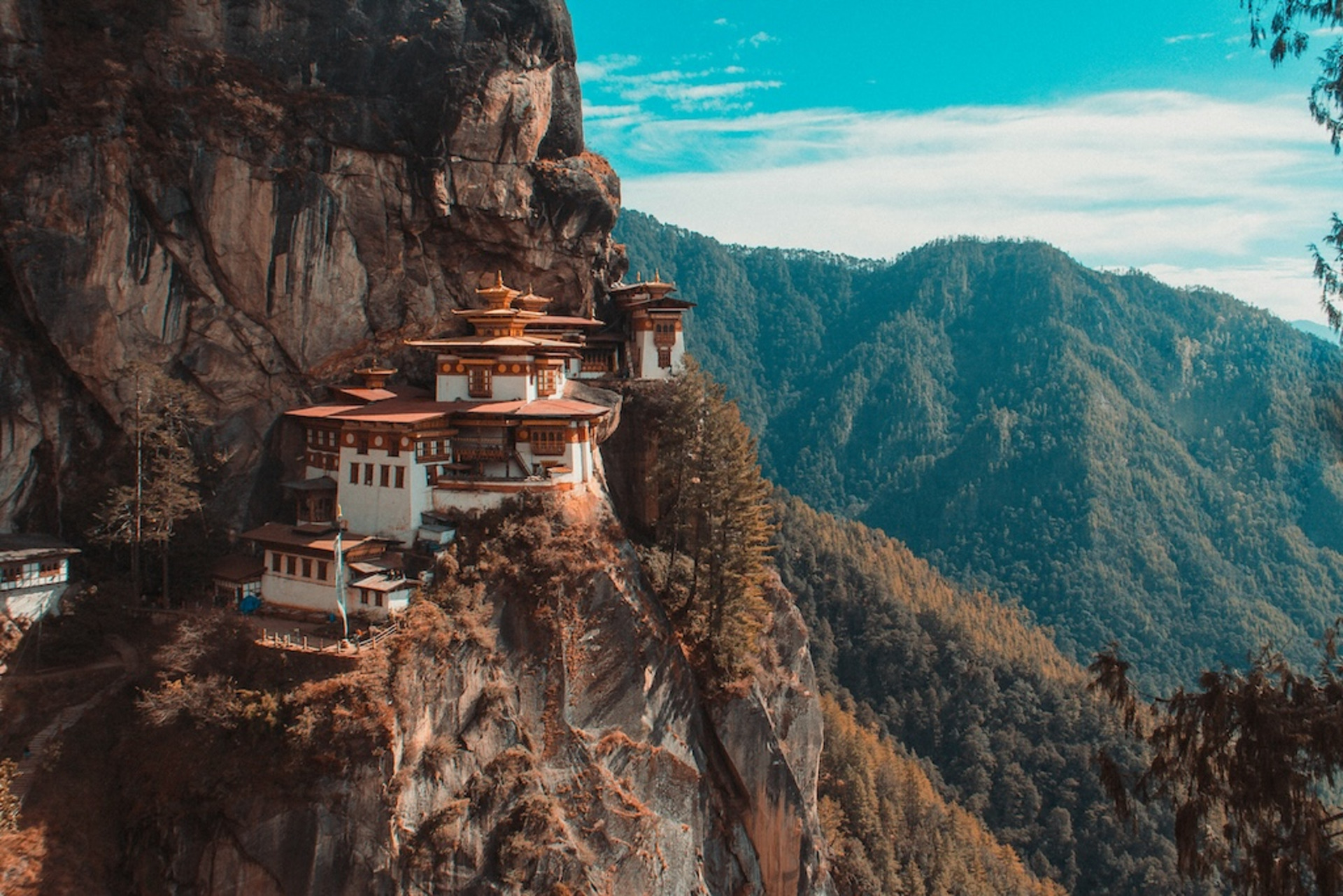 Das Taktshang-Kloster in Bhutan: Zu sehen sind eine Reihe von Tempeln, die sich an einen steilen Berghang schmiegen. Im Hintergrund sind weitere bewaldete Berghänge sichtbar.