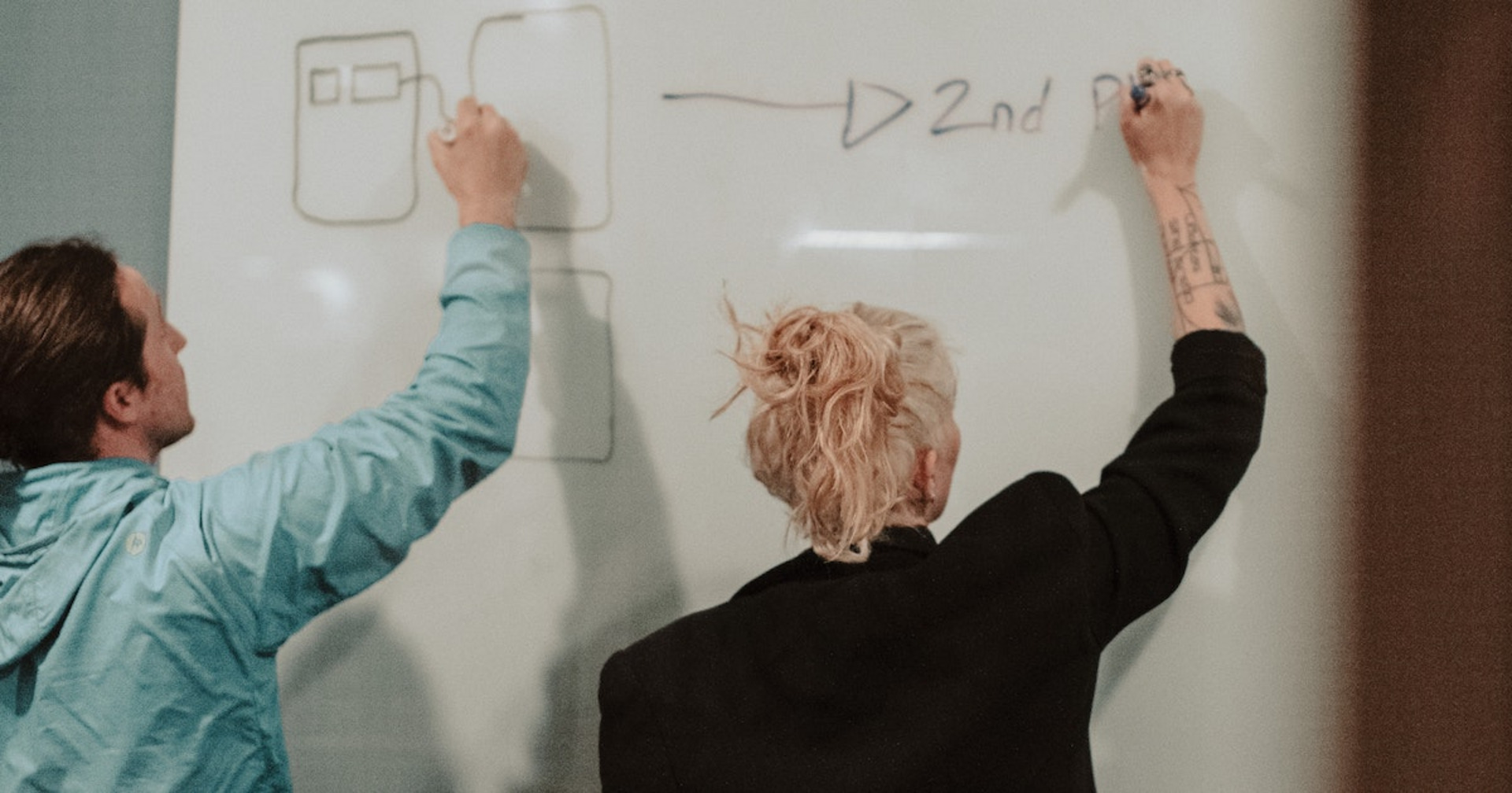 Zwei Personen schreiben auf einem Whiteboard mit dem Rücken zur Kamera.