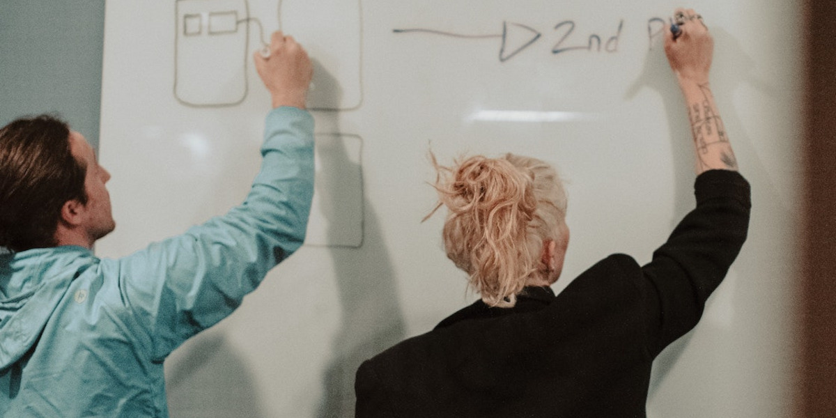Zwei Personen schreiben auf einem Whiteboard mit dem Rücken zur Kamera.