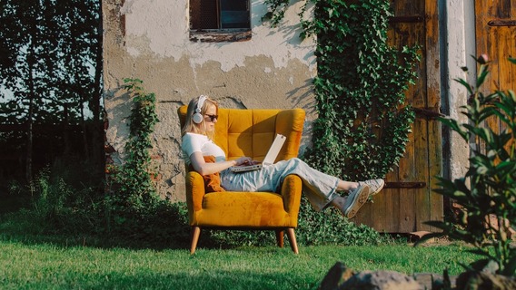 Eine junge Frau mit Sonnenbrille und weißen Kopfhörern sitzt draußen in einem gelben Sessel und tippt auf einem Laptop. Sie trägt ein weißes T-Shirt, eine helle Hose und Turnschuhe. Im Hintergrund ist ein altes Gebäude mit einer bewachsenen Holzfassade zu sehen.