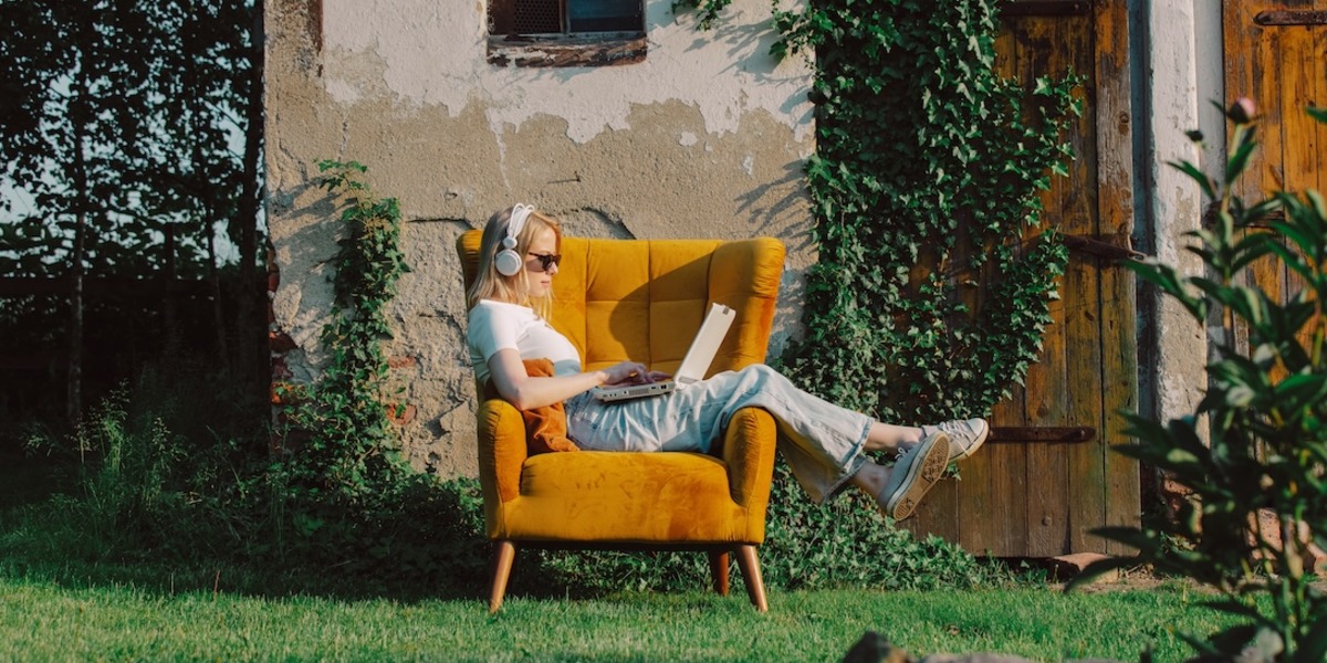 Eine junge Frau mit Sonnenbrille und weißen Kopfhörern sitzt draußen in einem gelben Sessel und tippt auf einem Laptop. Sie trägt ein weißes T-Shirt, eine helle Hose und Turnschuhe. Im Hintergrund ist ein altes Gebäude mit einer bewachsenen Holzfassade zu sehen.