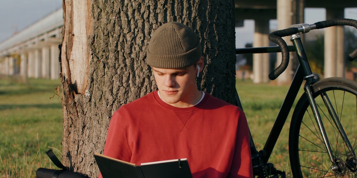 Ein junger Mann in rotem Pullover und Beanie-Mütze sitzt an einen Baum gelehnt auf einer Wiese und liest in einem schwarzen Notizbuch. Neben ihm liegen ein Rucksack und ein Kaffeebecher, im Hintergrund steht ein schwarzes Rennrad. 