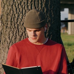 Ein junger Mann in rotem Pullover und Beanie-Mütze sitzt an einen Baum gelehnt auf einer Wiese und liest in einem schwarzen Notizbuch. Neben ihm liegen ein Rucksack und ein Kaffeebecher, im Hintergrund steht ein schwarzes Rennrad. 