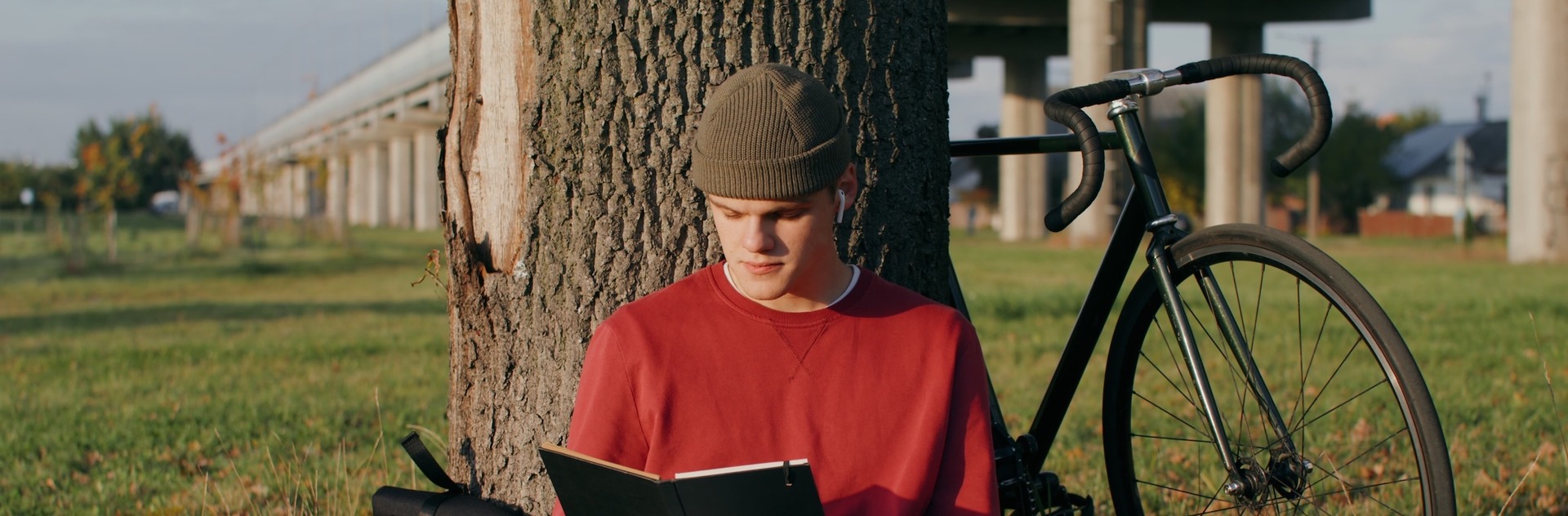 Ein junger Mann in rotem Pullover und Beanie-Mütze sitzt an einen Baum gelehnt auf einer Wiese und liest in einem schwarzen Notizbuch. Neben ihm liegen ein Rucksack und ein Kaffeebecher, im Hintergrund steht ein schwarzes Rennrad. 